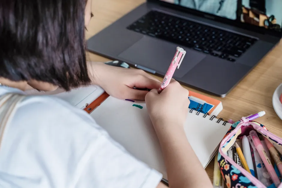 A young girl engaged in distance learning, using a laptop and taking notes in a notebook at home.