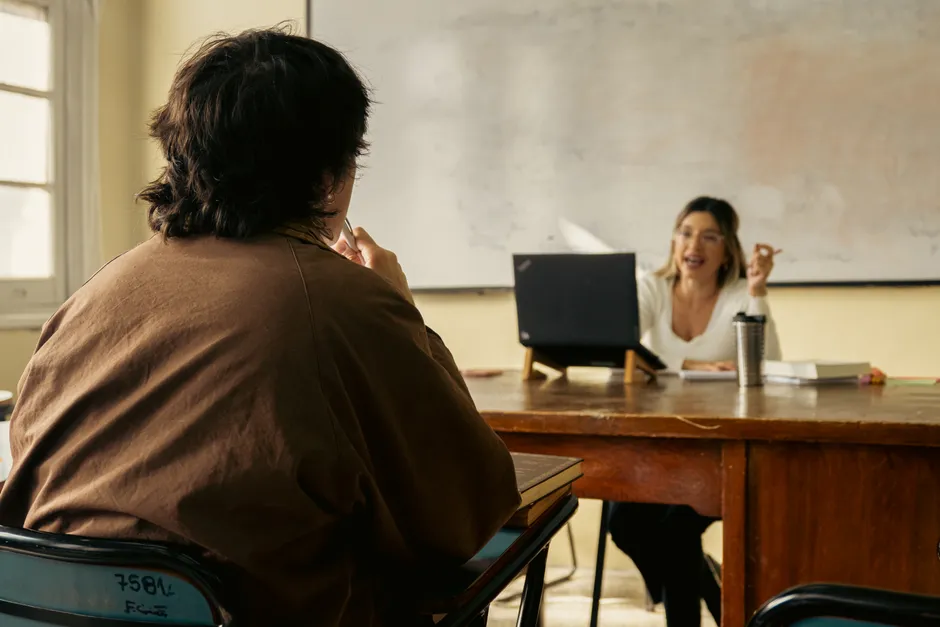 Student and teacher engage in discussion in a classroom setting with a laptop and whiteboard visible.