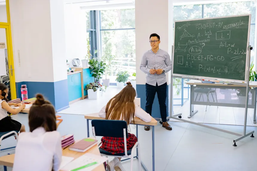 Teacher instructing students in a bright classroom with a blackboard filled with math equations.