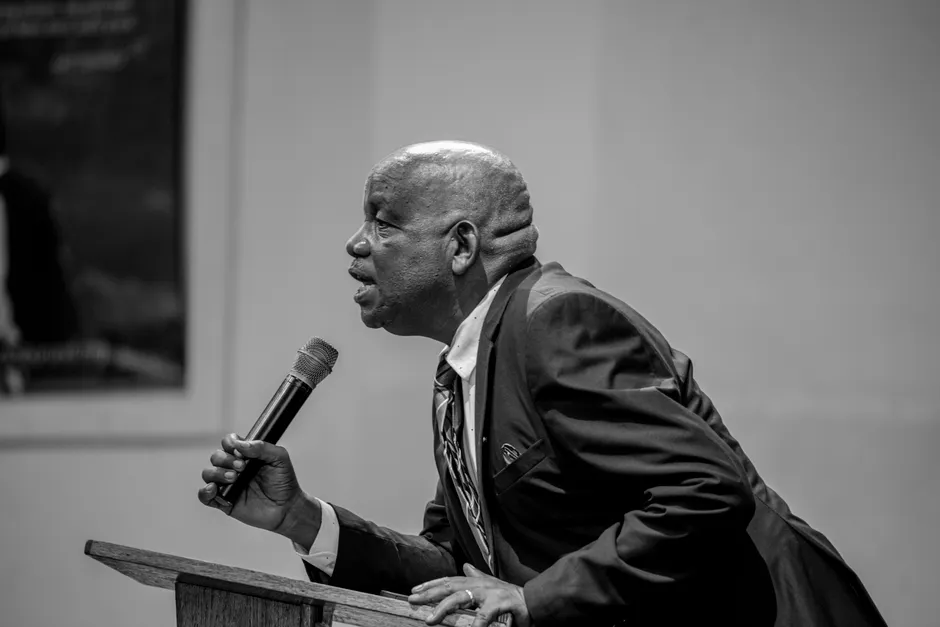 Black and white photo of a speaker at a podium with a microphone, passionately engaging an audience.