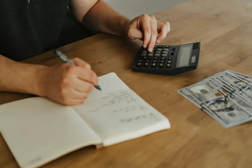 Close-up of hands using a calculator with notebook and cash on a wooden table.