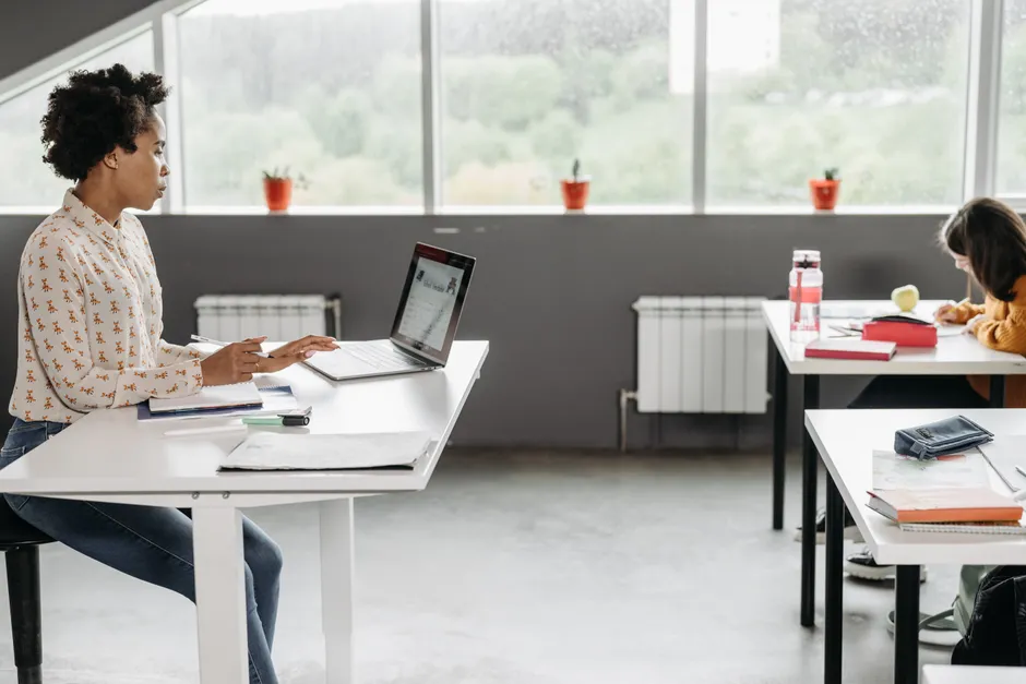 A teacher focused on her laptop in a bright, modern classroom setting.
