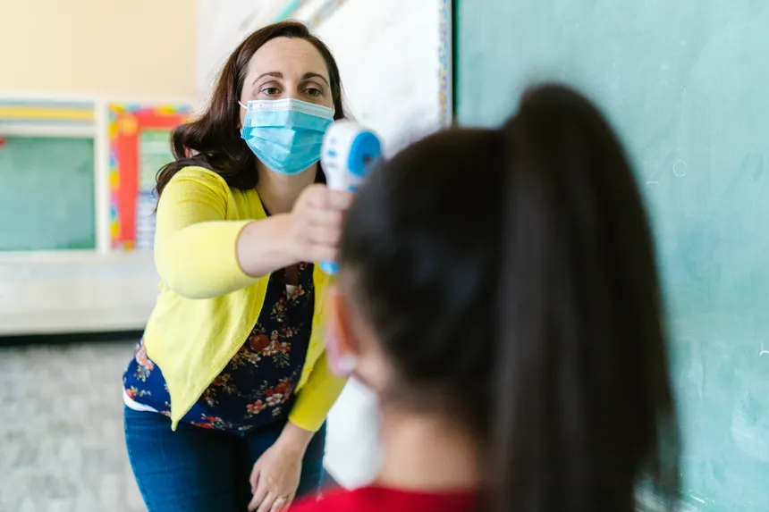 Female teacher checks student's temperature with thermometer in classroom.