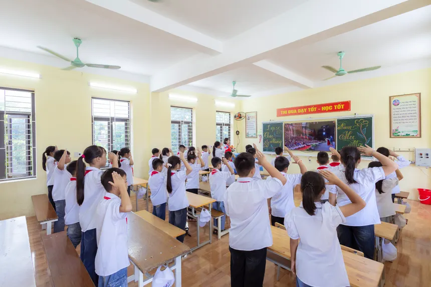 Students in uniforms saluting in a bright classroom during a school session.
