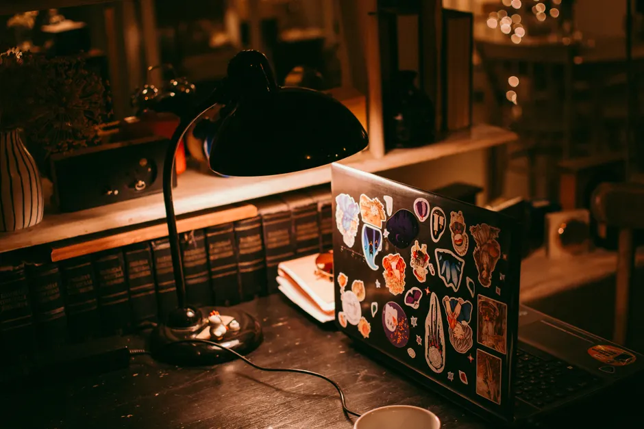 Warm lighting illuminates a workspace with a laptop, lamp, and books.