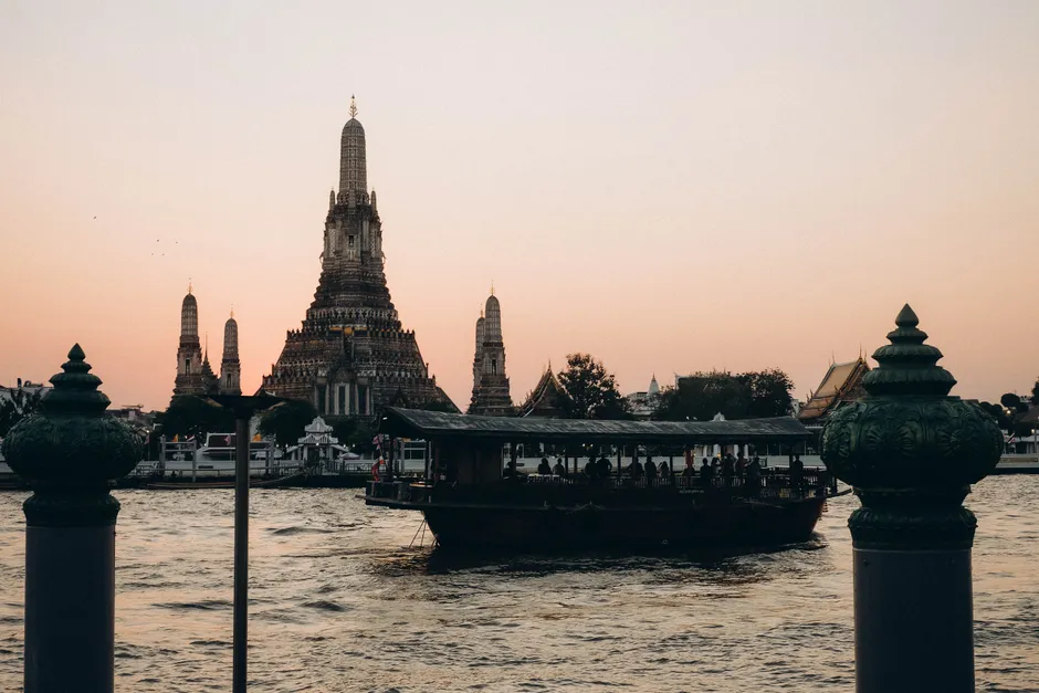 A serene view of Wat Arun Temple with a ferry on the Chao Phraya River at sunset in Bangkok.